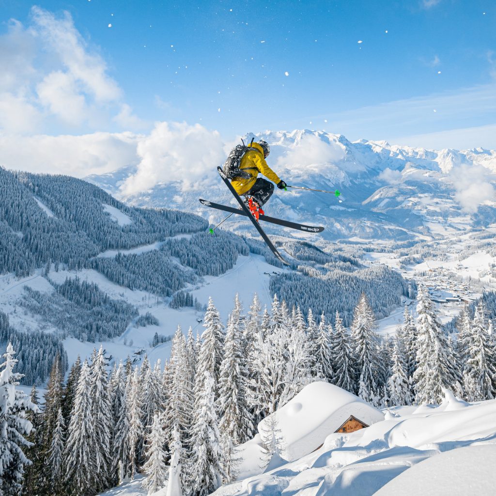 Ein Skifahrer in einer gelben Jacke springt mitten in der Luft über eine verschneite Berglandschaft mit Kiefern.