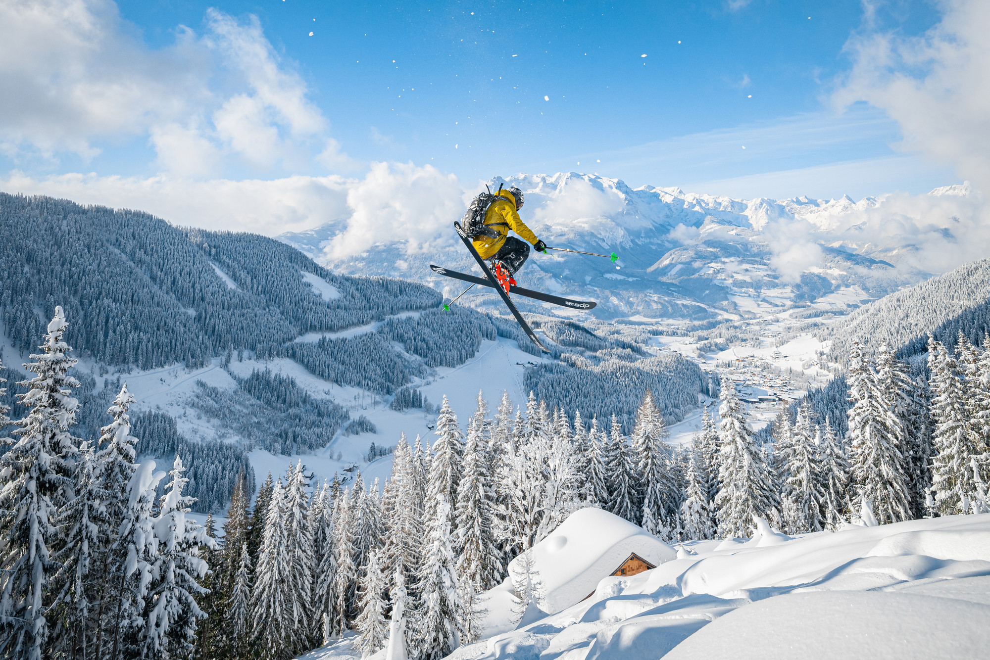 Ein Skifahrer in einer gelben Jacke springt mitten in der Luft über eine verschneite Berglandschaft mit Kiefern.