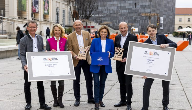 Sechs Personen stehen auf einem Stadtplatz mit Urkunden und Plaketten in der Hand und lächeln in die Kamera.