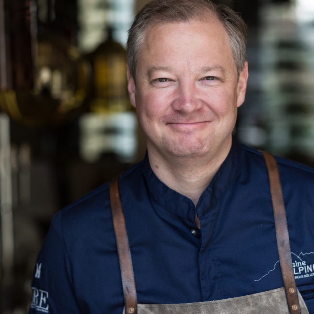 Ein lächelnder Koch in einer marineblauen Jacke und einer braunen Schürze steht in einer Restaurantküche.