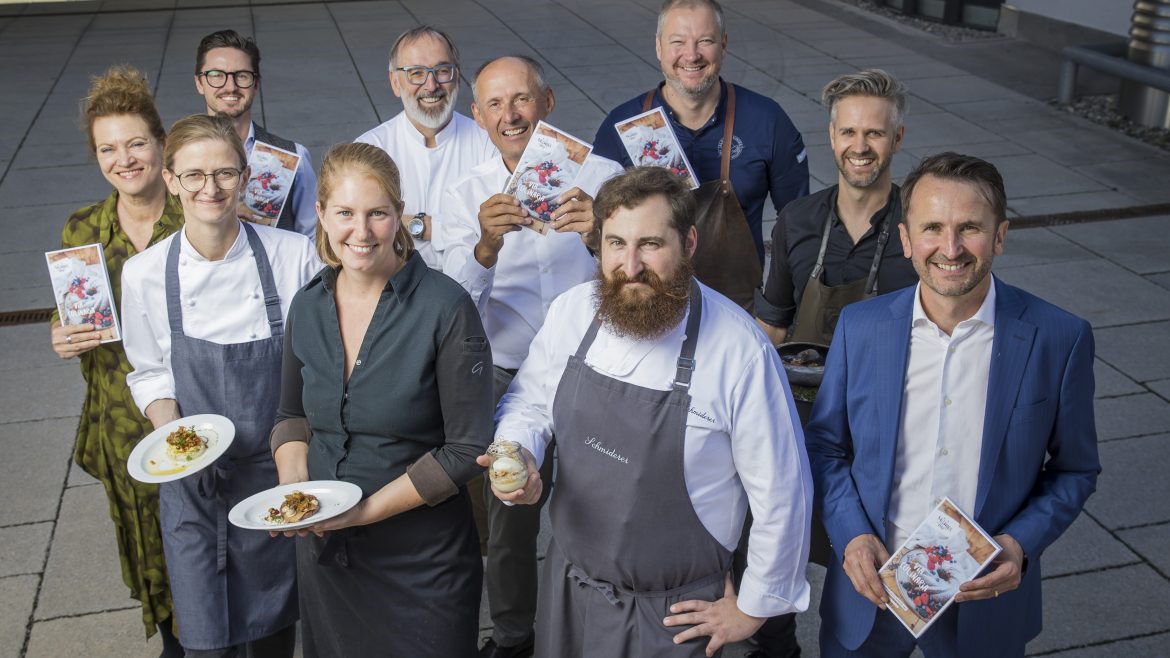 Eine Gruppe von lächelnden Köchen und Fachleuten posiert im Freien, einige halten Gerichte, andere Broschüren in der Hand.