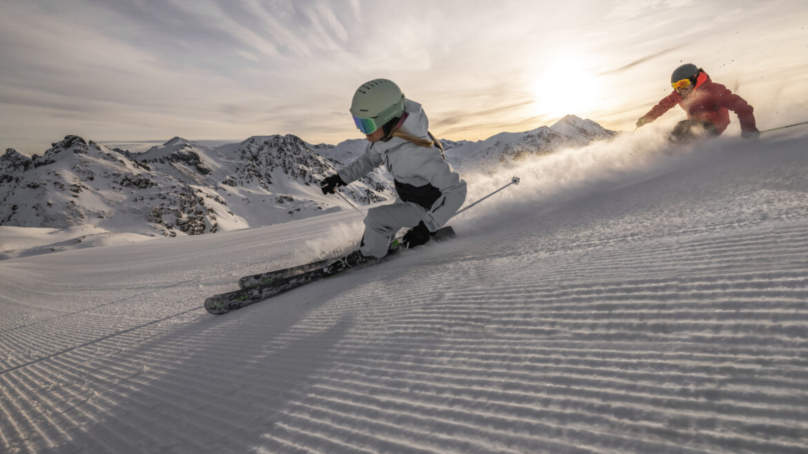 Zwei Skifahrer fahren auf einem verschneiten Berg mit Sonne und schroffen Gipfeln im Hintergrund bergab.