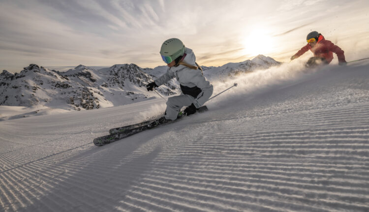 Zwei Skifahrer fahren auf einem verschneiten Berg mit Sonne und schroffen Gipfeln im Hintergrund bergab.
