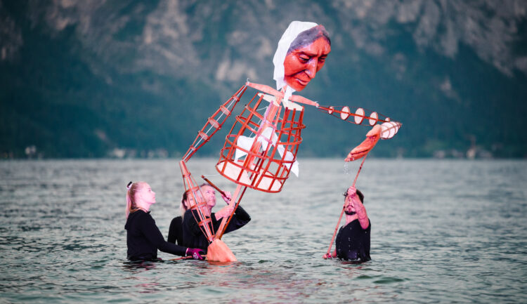 Drei Personen im Wasser bedienen eine große Puppe mit bemaltem Gesicht und Holzrahmen, im Hintergrund Berge.