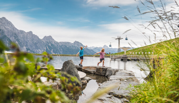 Zwei Kinder spielen auf Felsen neben einem Bergsee, mit Seilbahnen und Bergen im Hintergrund.