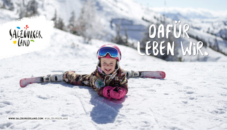Lächelndes Kind in Skikleidung auf einer verschneiten Piste mit Skiern, vor einem Berghintergrund.