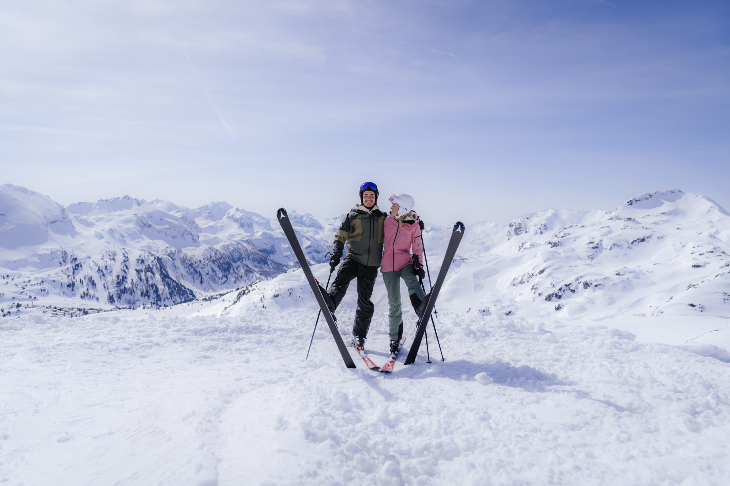Zwei Personen in Skiausrüstung stehen mit gekreuzten Skiern auf einem verschneiten Berg, umgeben von verschneiten Gipfeln.