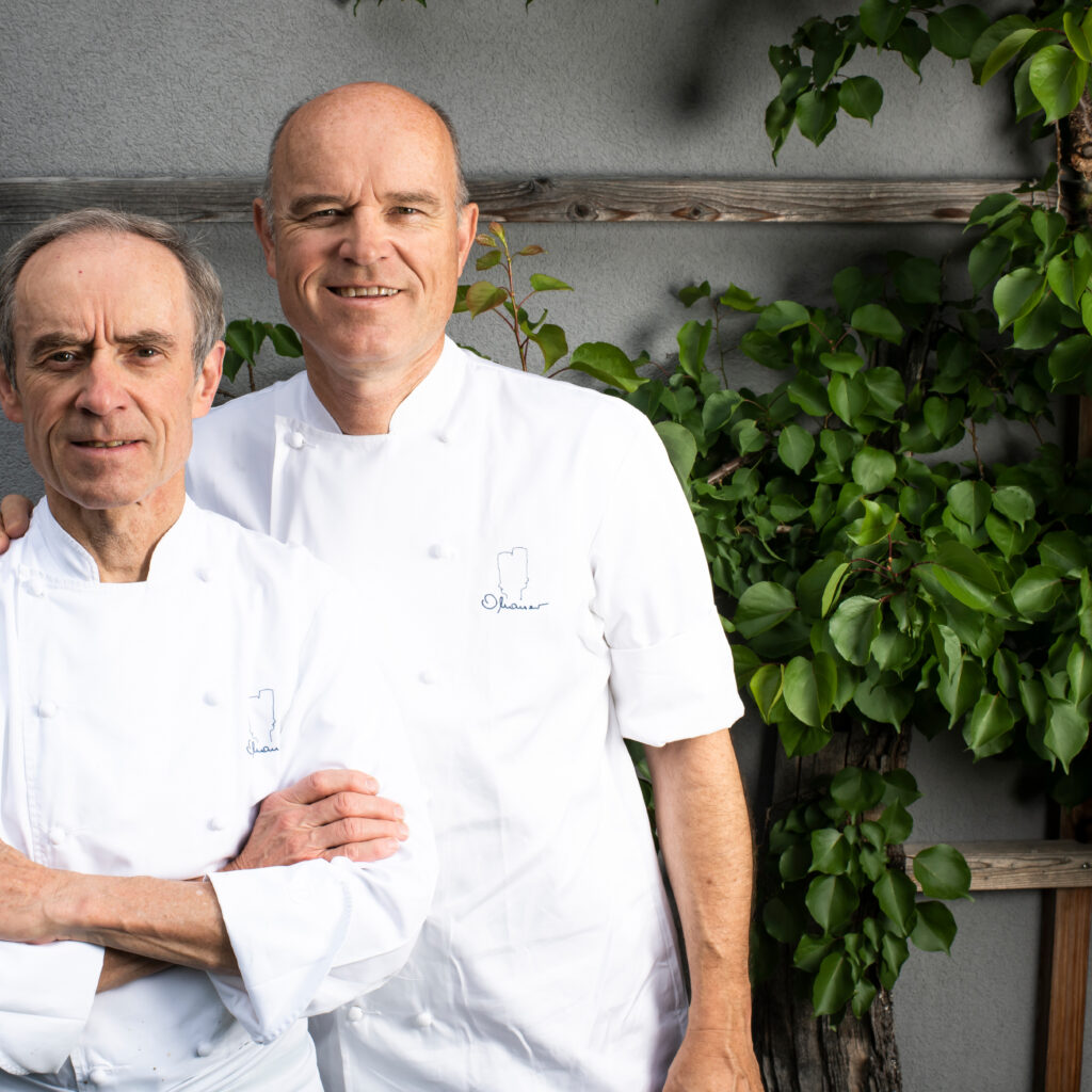 Zwei Köche in weißen Uniformen stehen draußen vor einer grünen Pflanze und einem Holzspalier zusammen.