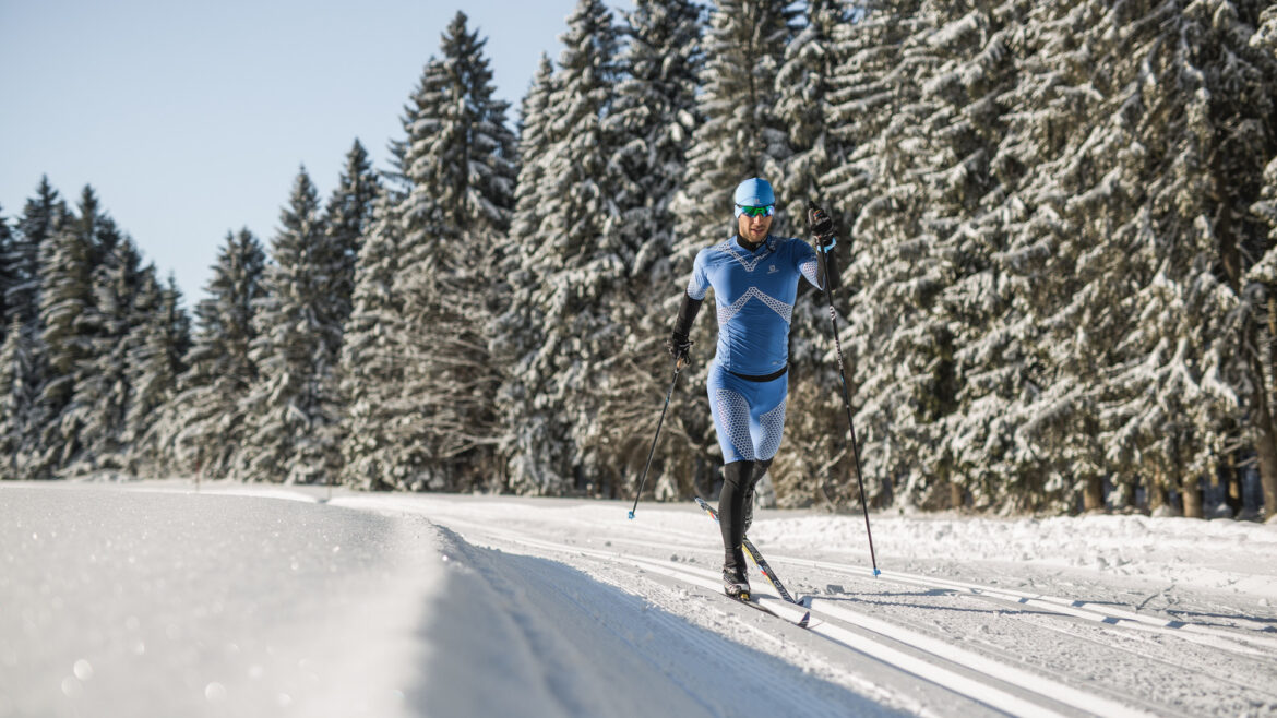 Ein Skifahrer in blauer Ausrüstung fährt Langlaufski auf einer verschneiten Loipe neben schneebedeckten Bäumen unter einem klaren Himmel.
