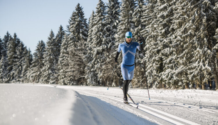Ein Skifahrer in blauer Ausrüstung fährt Langlaufski auf einer verschneiten Loipe neben schneebedeckten Bäumen unter einem klaren Himmel.