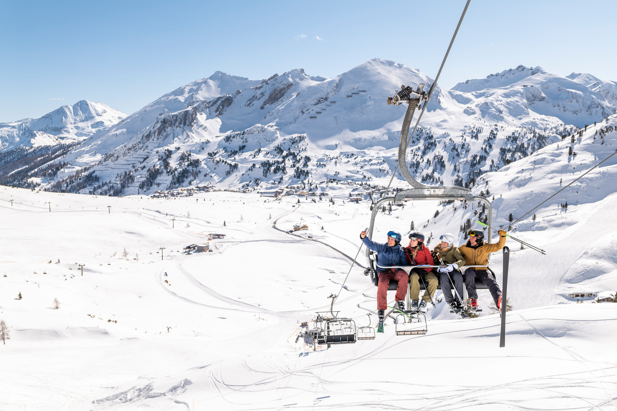 Drei Skifahrer fahren an einem klaren, sonnigen Tag mit einem Lift über eine verschneite Berglandschaft.
