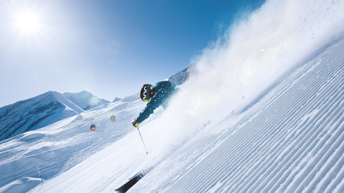 Ein Skifahrer rast an einem sonnigen Tag einen verschneiten Berghang hinunter und wirbelt Pulverschnee auf, während im Hintergrund Skilifte zu sehen sind.