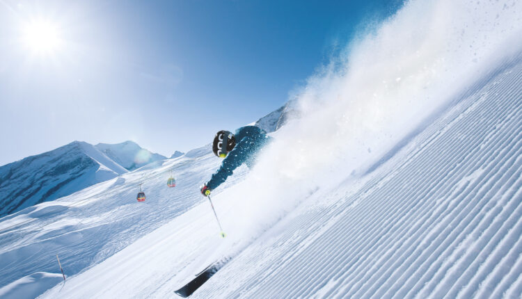Ein Skifahrer rast an einem sonnigen Tag einen verschneiten Berghang hinunter und wirbelt Pulverschnee auf, während im Hintergrund Skilifte zu sehen sind.