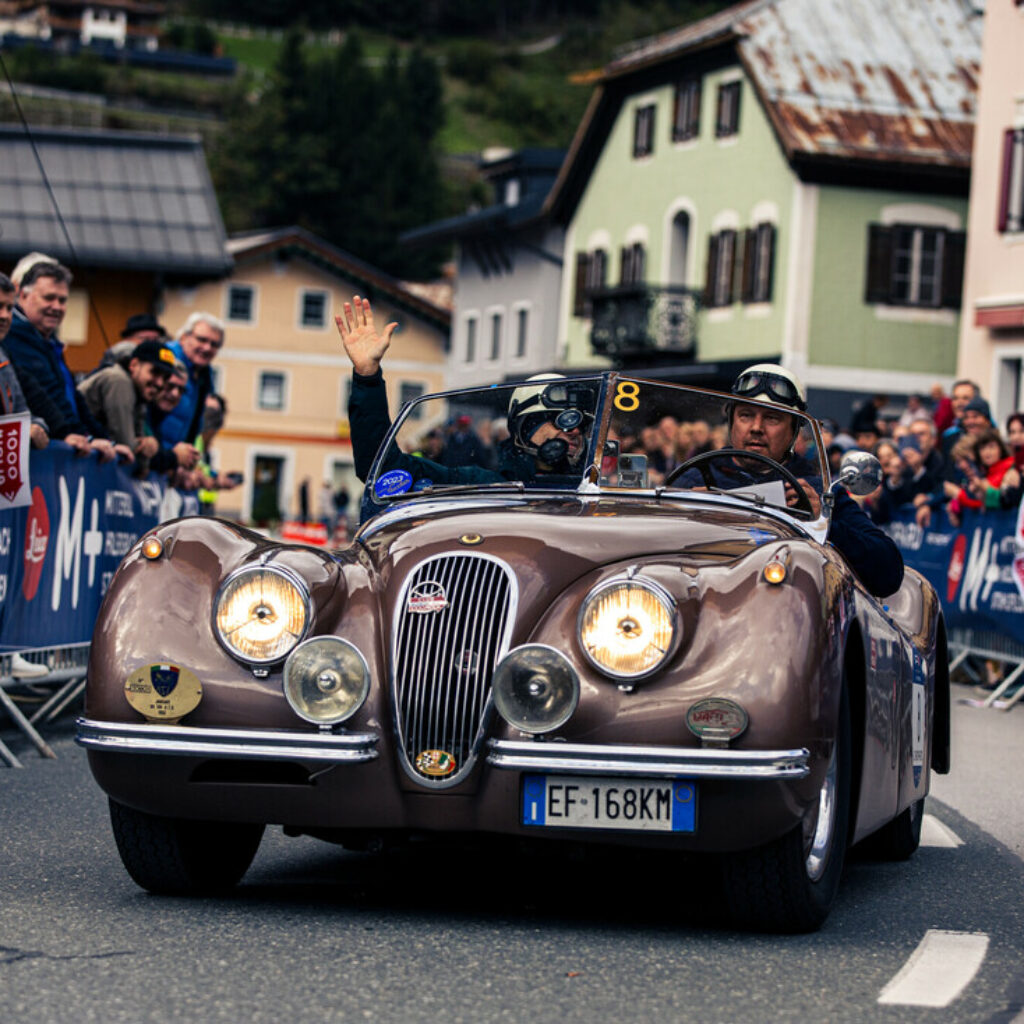 Ein klassisches braunes Cabriolet mit zwei Personen fährt an einer jubelnden Menschenmenge auf einer Straßenveranstaltung vorbei.