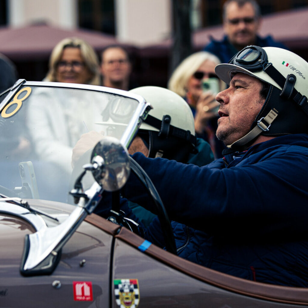 Ein Mann in historischer Rennkleidung fährt einen Oldtimer vor einer Menschenmenge bei einer Veranstaltung im Freien.