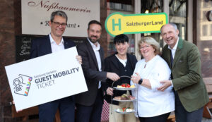 Fünf Personen lächeln vor einem Restaurant, in der Hand ein Schild, ein Tablett mit Süßspeisen und ein Salzburger Tourismusschild.