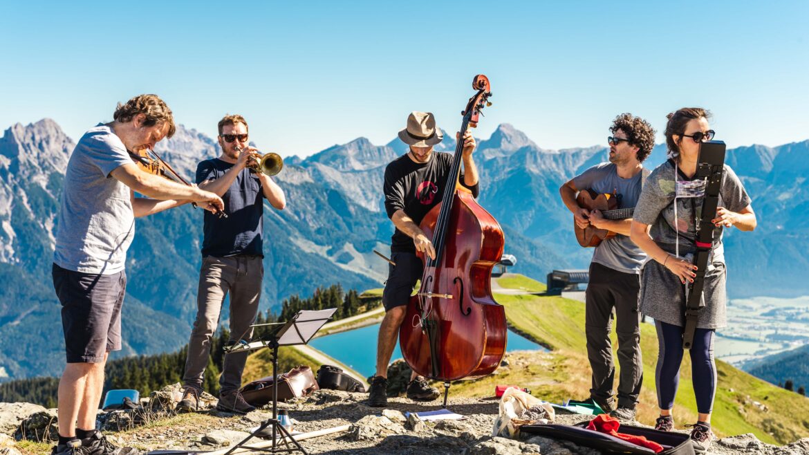Fünf Musiker spielen Instrumente im Freien auf einem Berg mit blauem Himmel und malerischen Gipfeln im Hintergrund.