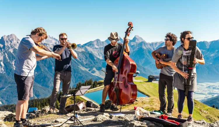 Fünf Musiker spielen Instrumente im Freien auf einem Berg mit blauem Himmel und malerischen Gipfeln im Hintergrund.