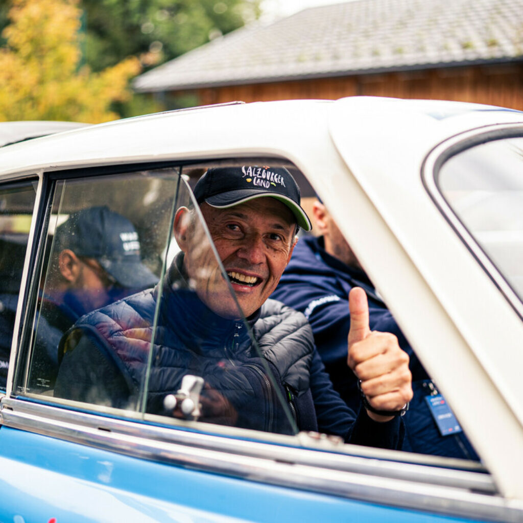 Ein lächelnder Mann in einem Auto zeigt den Daumen nach oben, er trägt eine Mütze und eine Jacke, draußen stehen Bäume und ein Gebäude.