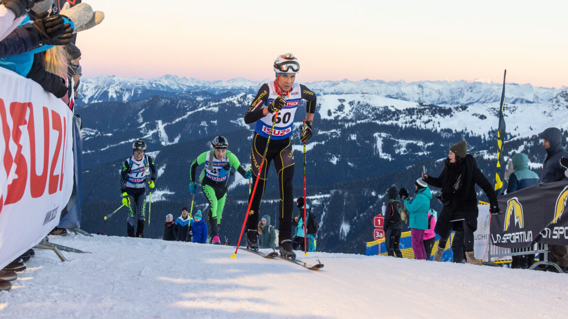 Skifahrer, die auf einem verschneiten Berg bergauf fahren, mit Zuschauern und alpiner Landschaft im Hintergrund.
