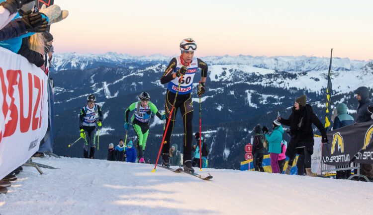 Skifahrer, die auf einem verschneiten Berg bergauf fahren, mit Zuschauern und alpiner Landschaft im Hintergrund.