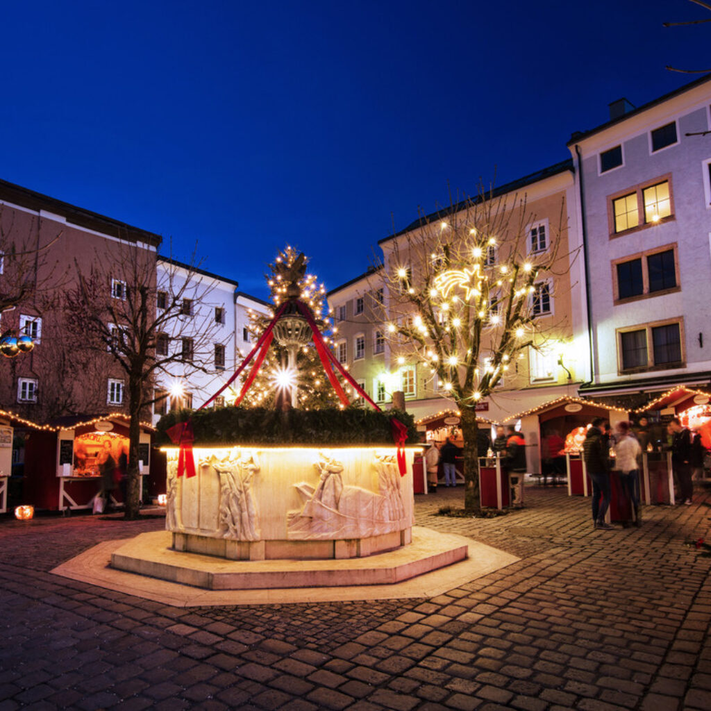 Ein geschmückter Springbrunnen, umgeben von kleinen Marktständen und beleuchteten Bäumen auf einem Platz in der Abenddämmerung.