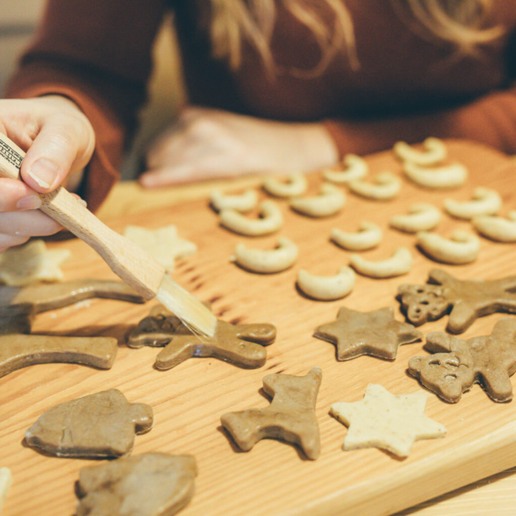 Eine Hand bestreicht mit einem hölzernen Backpinsel rohe Weihnachtsplätzchen-Teigformen auf einem hölzernen Schneidebrett mit Glasur.