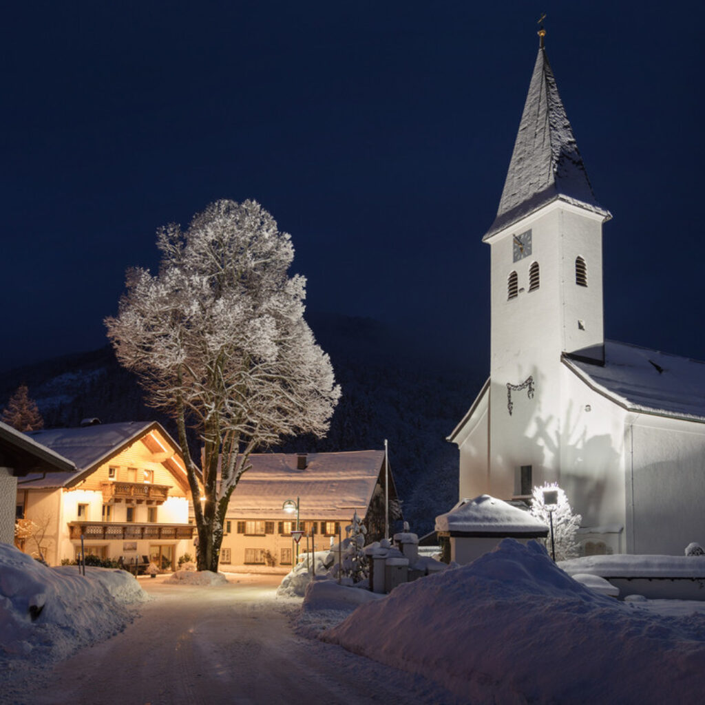 Ein schneebedeckter Dorfplatz bei Nacht mit einer weißen Kirche und traditionellen Häusern.