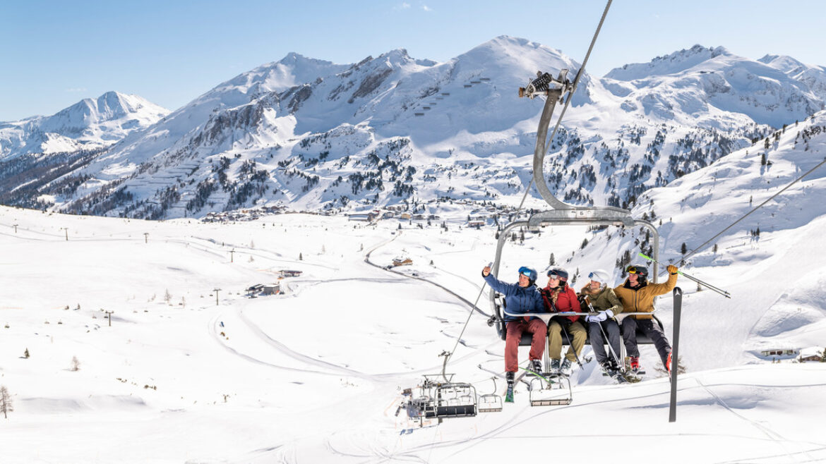 Vier Personen fahren an einem sonnigen Tag mit einem Skilift über verschneite Berge, umgeben von einer Winterlandschaft.