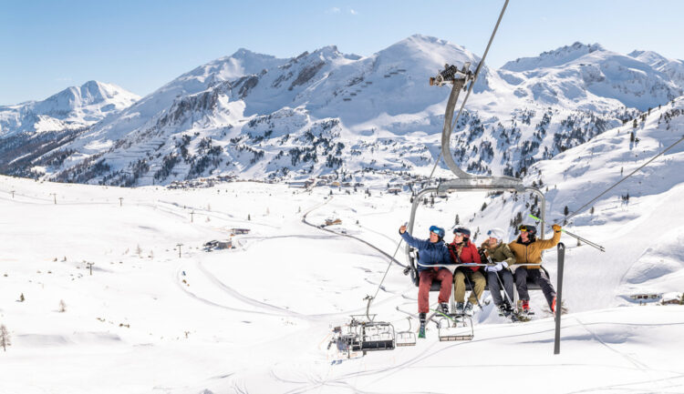 Vier Personen fahren an einem sonnigen Tag mit einem Skilift über verschneite Berge, umgeben von einer Winterlandschaft.