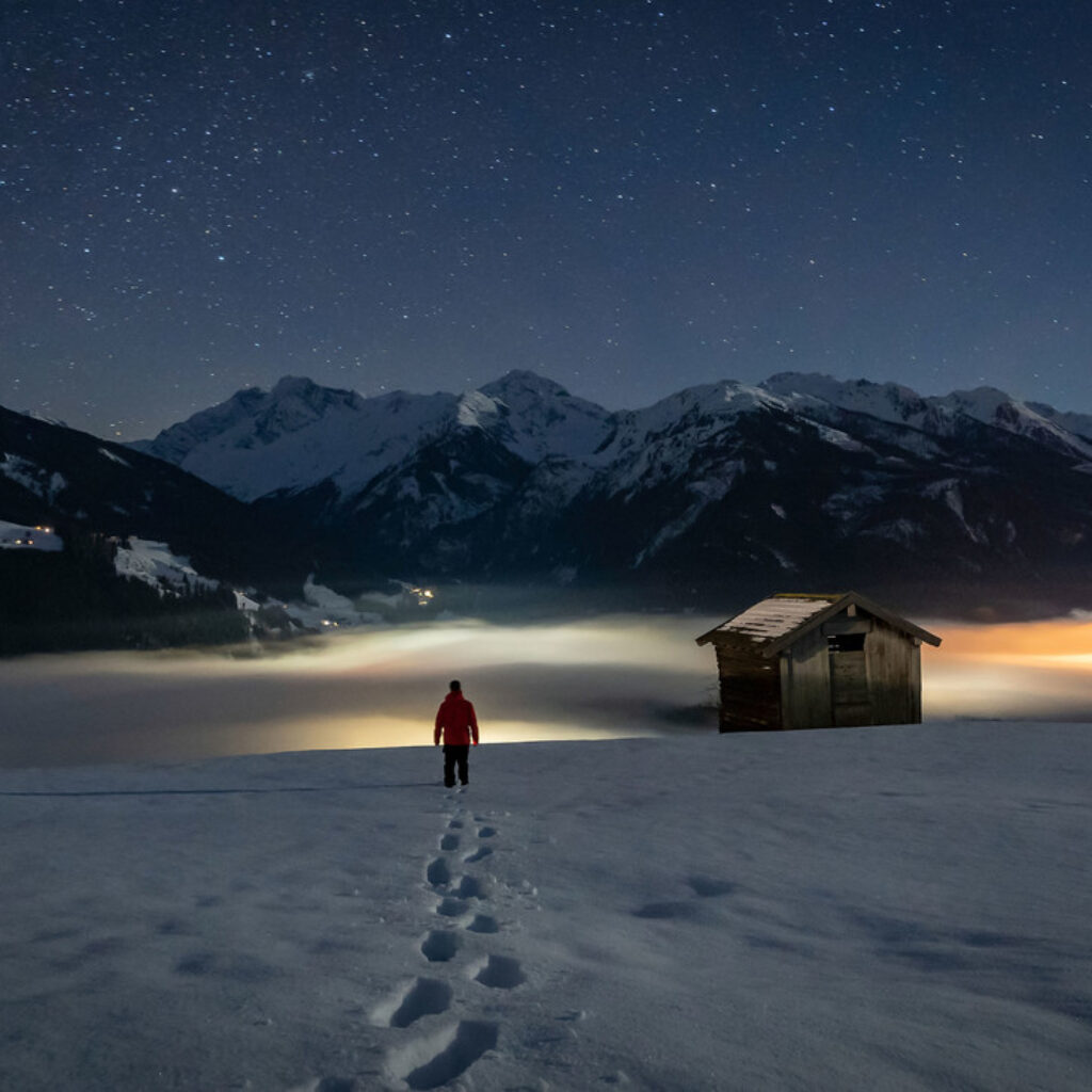 Eine Person in einer roten Jacke steht nachts vor einer österreichischen Berglandschaft im Schnee unter einem Sternenhimmel.