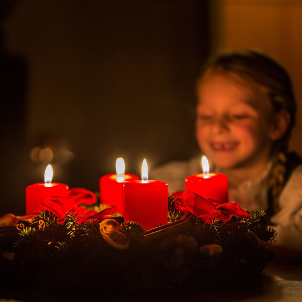 Ein Kind in traditioneller österreichischer Tracht betrachtet die vier brennenden Kerzen eines Adventskranzes.