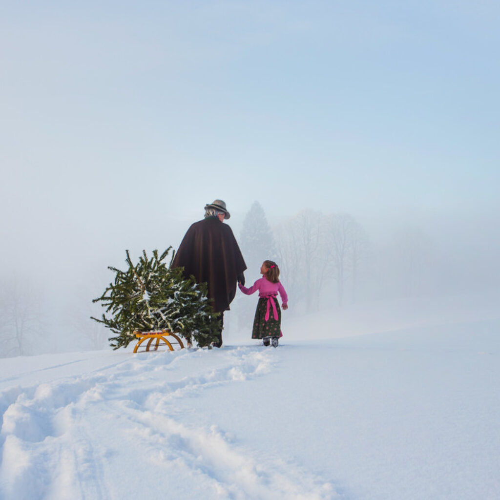 Eine Person und ein Kind in traditioneller österreichischer Tracht ziehen einen Weihnachtsbaum auf einem Schlitten durch eine verschneite Landschaft.