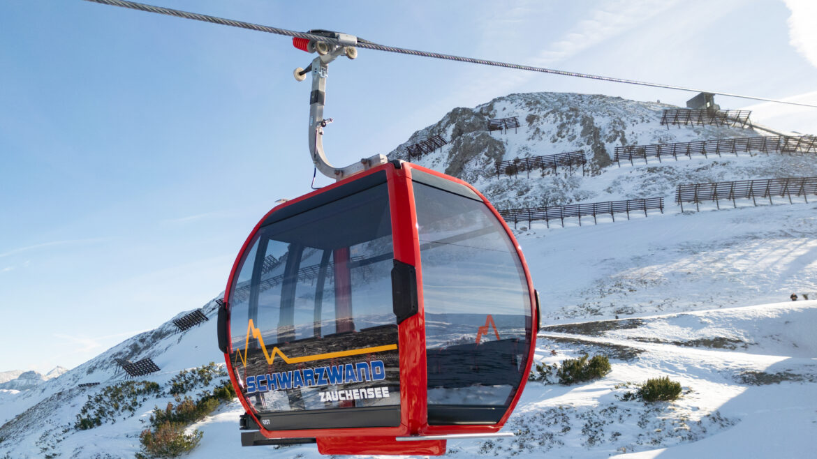 Rote Skigondel, die über einer verschneiten Berglandschaft mit blauem Himmel im Hintergrund schwebt.