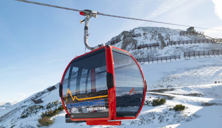 Rote Skigondel, die über einer verschneiten Berglandschaft mit blauem Himmel im Hintergrund schwebt.