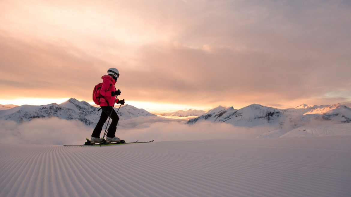 Person beim Skifahren bei Sonnenaufgang auf einer präparierten Piste mit schneebedeckten Bergen im Hintergrund.