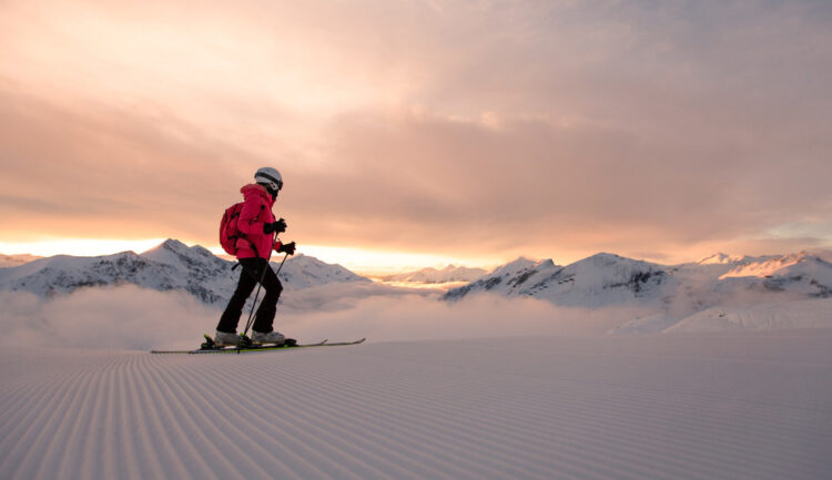 Person beim Skifahren bei Sonnenaufgang auf einer präparierten Piste mit schneebedeckten Bergen im Hintergrund.