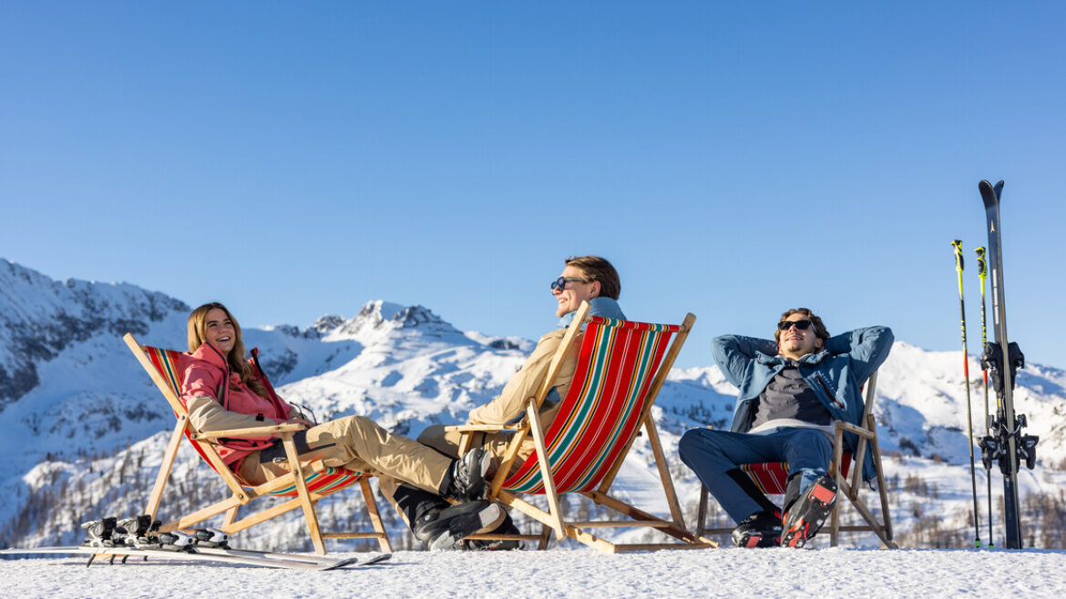 Drei Personen entspannen sich in Liegestühlen auf einem verschneiten Berg mit Skiern in der Nähe unter einem klaren blauen Himmel.
