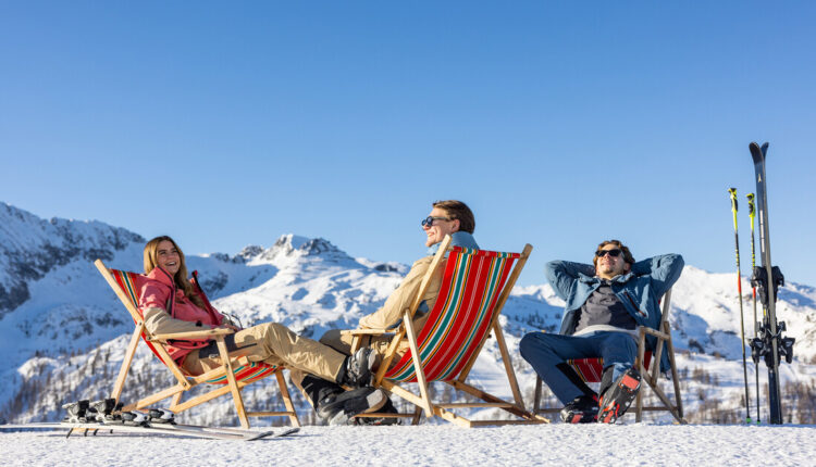 Drei Personen entspannen sich in Liegestühlen auf einem verschneiten Berg mit Skiern in der Nähe unter einem klaren blauen Himmel.