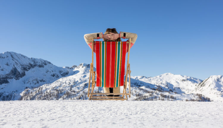 Person, die sich in einem gestreiften Liegestuhl auf verschneitem Boden entspannt, mit Blick auf schneebedeckte Berge unter einem klaren blauen Himmel.