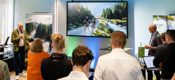 Eine Gruppe verfolgt den Vortrag eines Redners neben einem großen Bildschirm, der Radfahrer in einer Berglandschaft zeigt.