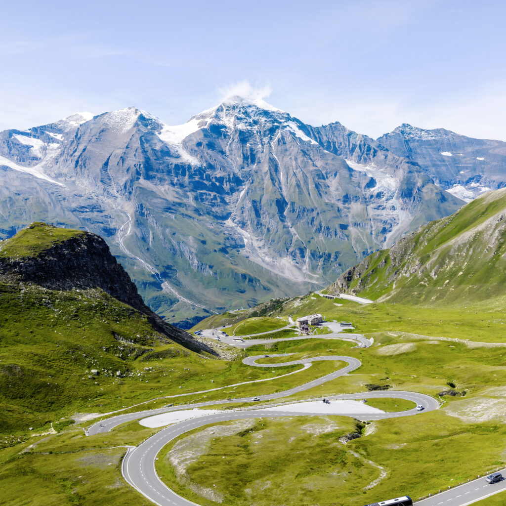 Eine kurvenreiche Bergstraße, die sich durch grüne Hügel mit schneebedeckten Gipfeln im Hintergrund unter einem klaren blauen Himmel schlängelt.