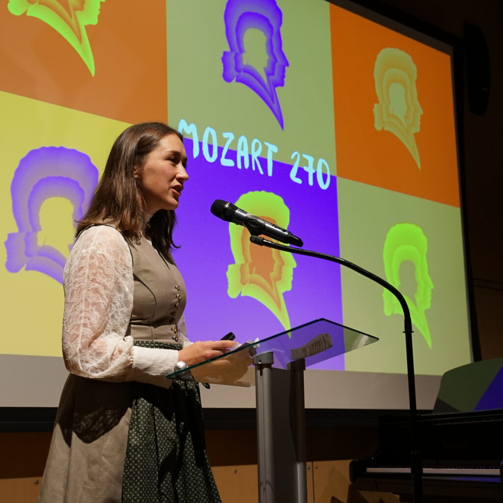 Eine Frau spricht auf einem Podium, während hinter ihr auf dem Bildschirm eine bunte Grafik von Mozart 270 angezeigt wird.