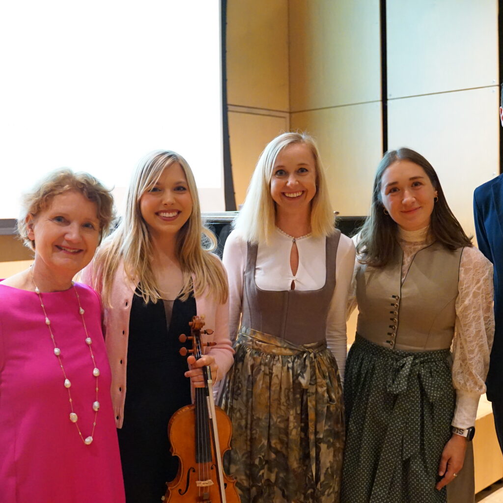 Fünf förmlich gekleidete Menschen stehen in einem Innenraum zusammen und lächeln, einer hält eine Geige.