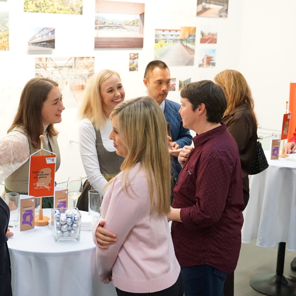 Menschen, die um kleine Tische herum stehen und sich unterhalten, bei einem Indoor-Networking oder einer gesellschaftlichen Veranstaltung mit Plakaten im Hintergrund.