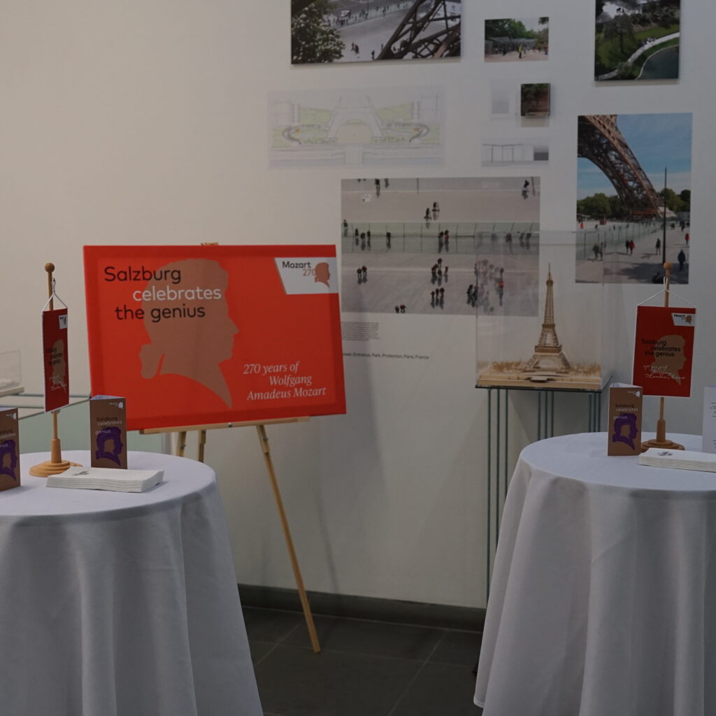 Zwei runde Tische mit Broschüren und Modellen vor architektonischen Displays und einem roten Veranstaltungsschild auf einer Staffelei.