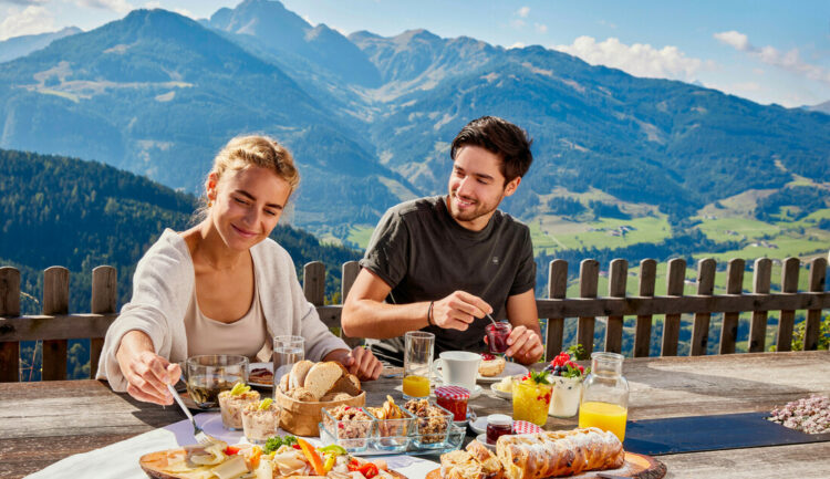 Ein lächelndes Paar genießt das Frühstück im Freien mit Bergen und grünen Tälern im Hintergrund.