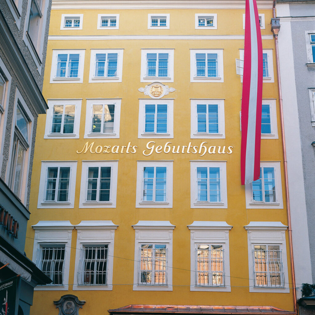 Gelbes Gebäude mit Mozarts Geburtshaus an der Fassade, großen Fenstern und einer herunterhängenden österreichischen Flagge.