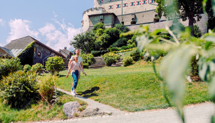 Ein Paar geht an einem sonnigen Tag einen Gartenweg hinauf in Richtung eines großen historischen Gebäudes.