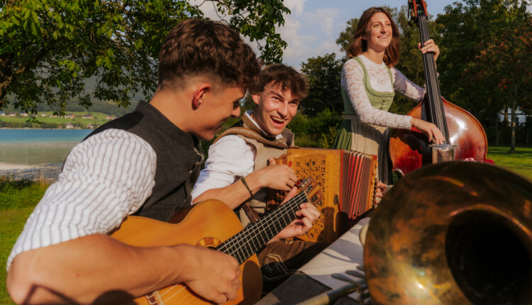 Drei junge Musiker spielen Gitarre, Akkordeon und Kontrabass an einem sonnigen Tag im Freien, lächeln und haben Spaß an der Musik.